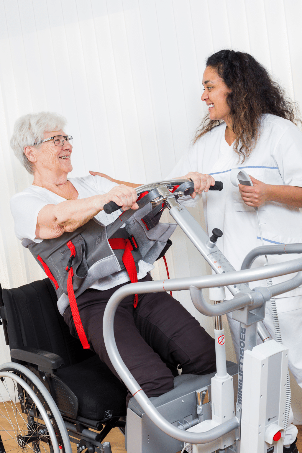 Woman with caretaker smiling while helping her out of a wheelchair using a sit to stand patient lift